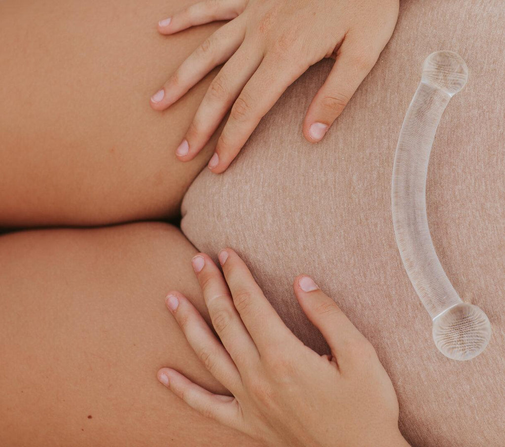 Curved clear glass pleasure wand with thicker spherical heads. Pictured held by model on lower torso. Model is wearing beige coloured underwear.